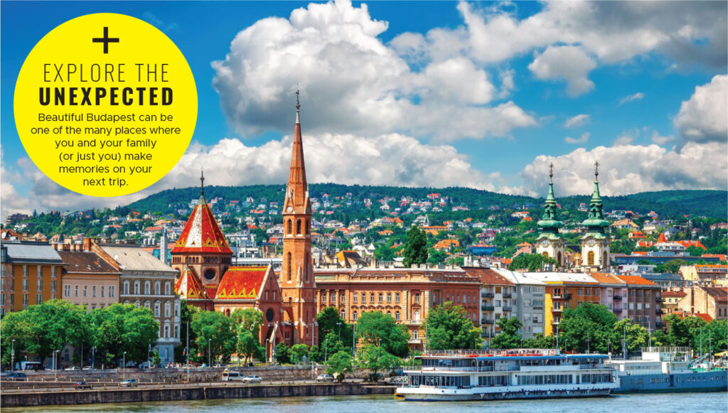 View of Budapest cityscape with colorful rooftops, church spires, green hills in the background, and a large boat docked on the river under a partly cloudy sky.