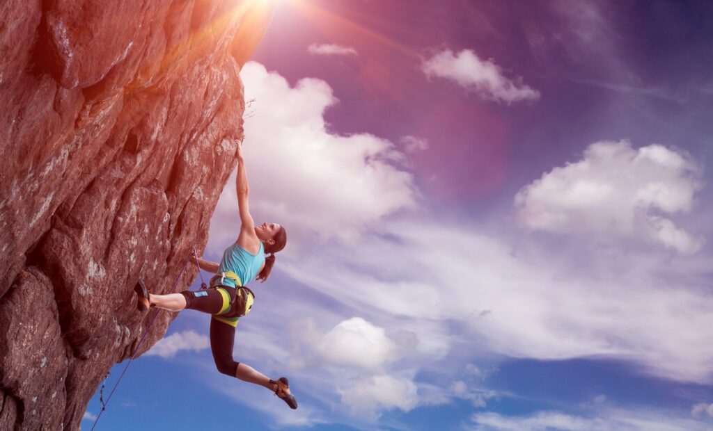Climber wearing a safety harness ascending a steep rock face beneath a cloudy sky.
