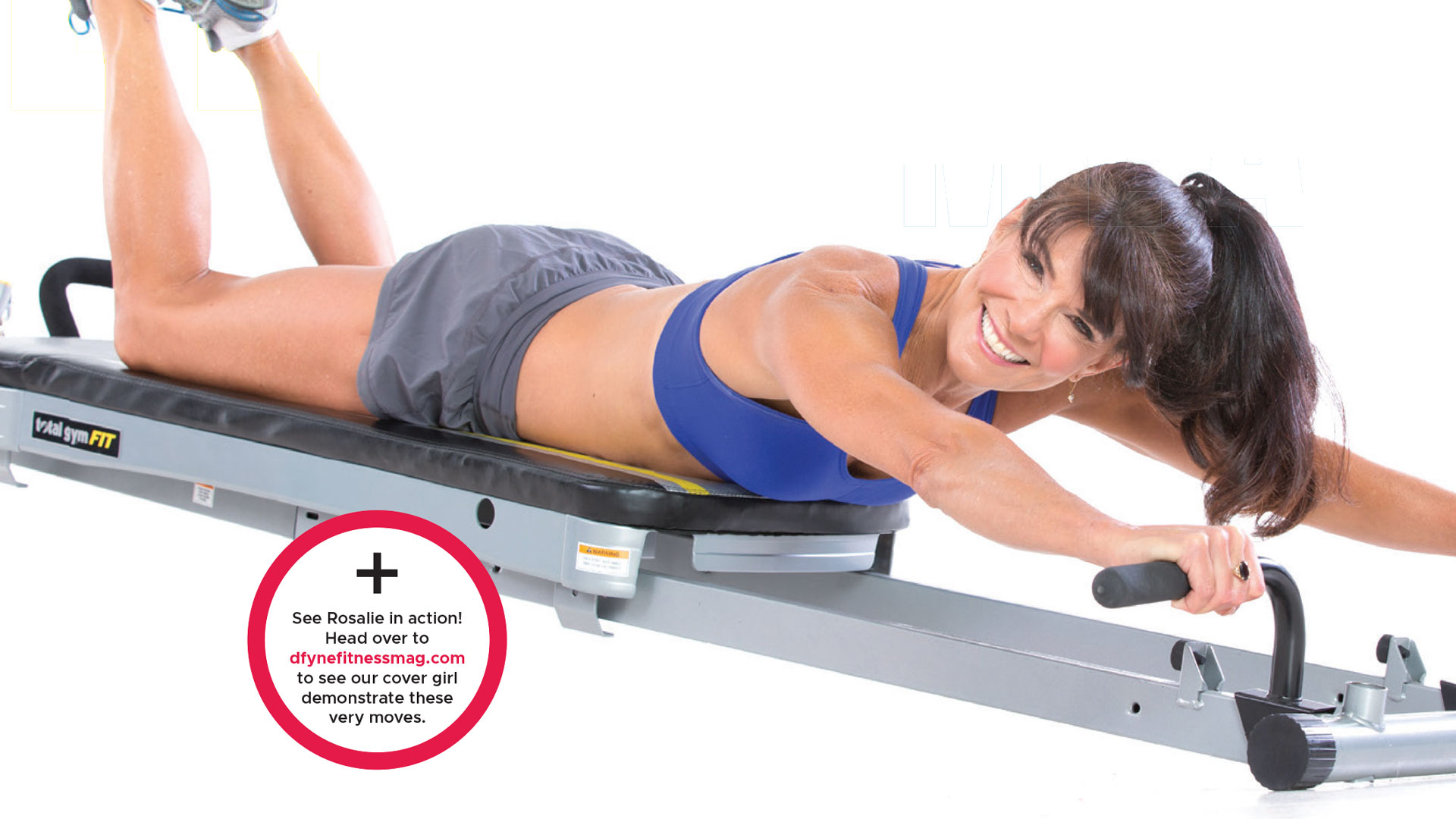 Woman in blue sports bra and gray shorts lying face down on a fitness machine, smiling and holding handles with arms extended forward.
