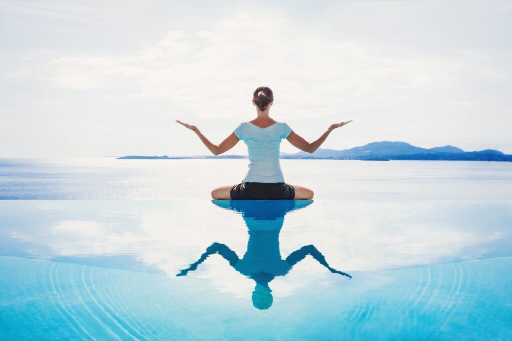 Woman sitting cross-legged at the edge of a pool with her reflection in the water.