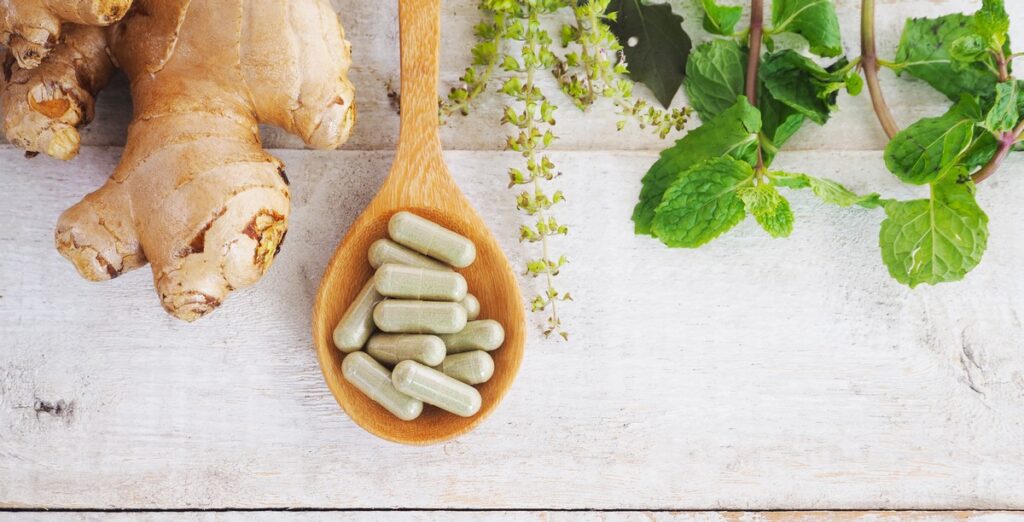 Capsules in a wooden spoon beside ginger and fresh herbs.