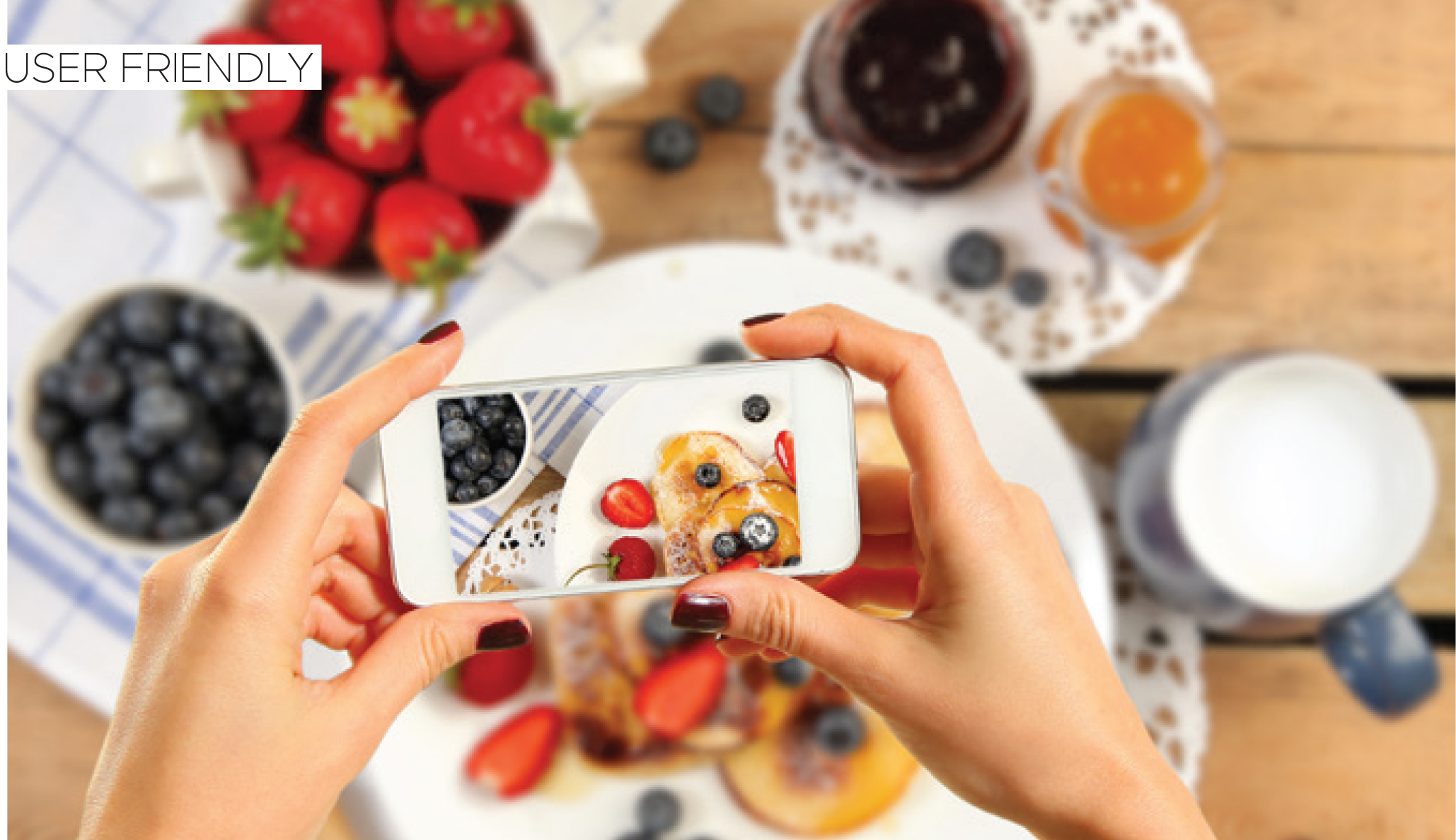 Hands holding a smartphone to photograph a breakfast spread with fruit and drinks.