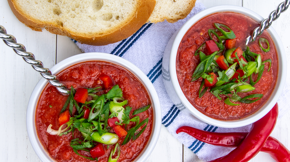 Two bowls of red soup topped with chopped vegetables and herbs, served with bread.