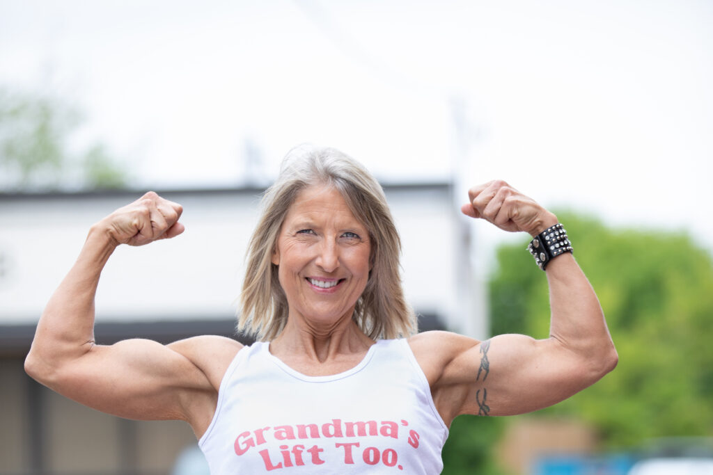 Smiling woman flexing both arms in a white tank top reading “Grandmas Lift Too”.