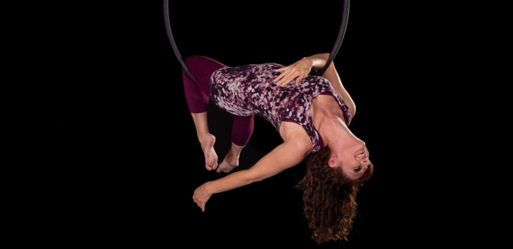 Woman performing an inverted pose in an aerial hoop against a black background.