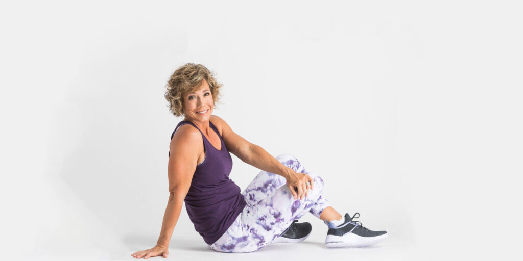 Woman in a purple workout top and patterned leggings seated on the floor.