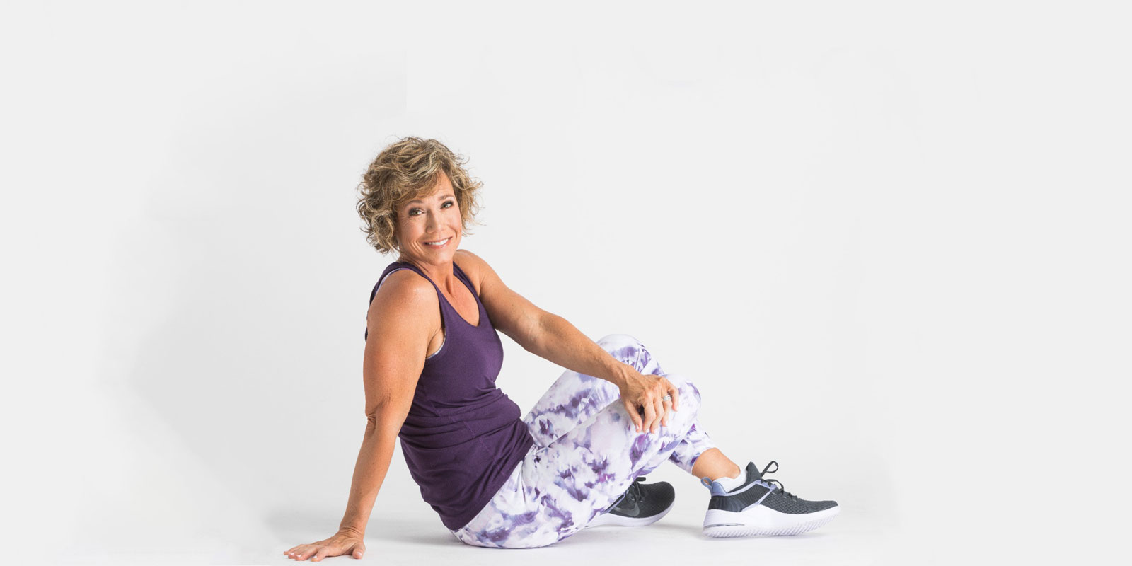 Woman in a purple workout top and patterned leggings seated on the floor.