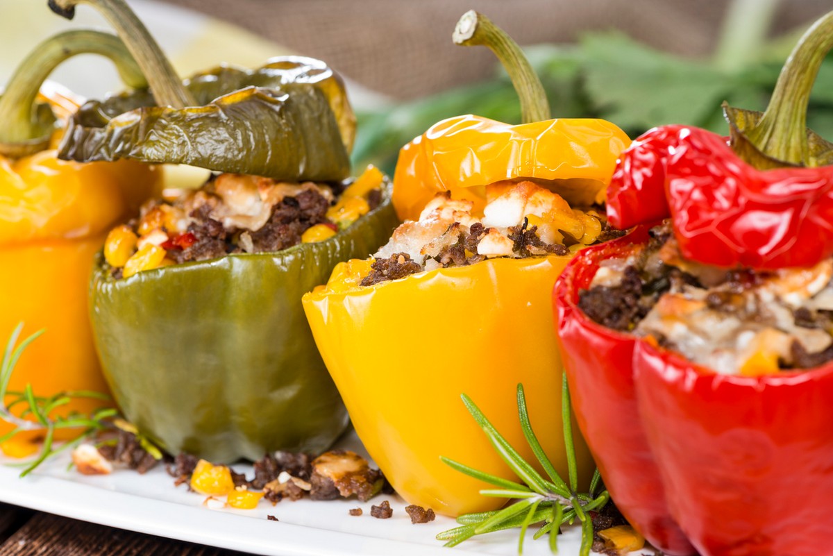 Close-up of green, yellow, and red bell peppers stuffed with a mixture of ground meat, corn, and cheese on a white plate with rosemary garnish.