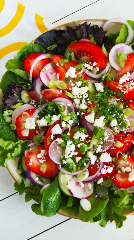 Salad with mixed baby greens, sliced strawberries, radishes, red onion, crumbled white cheese, and chopped green herbs in a bowl.