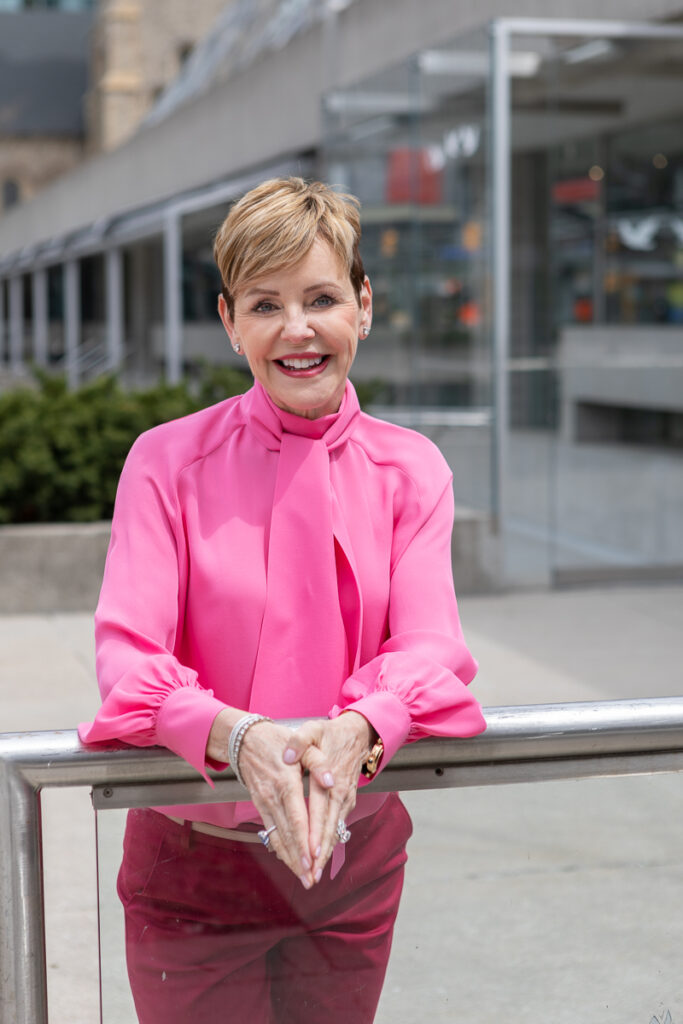 Smiling woman with short hair wearing a bright pink blouse outside a modern building.