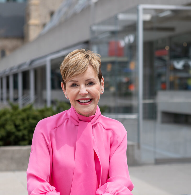 Smiling woman with short hair wearing a bright pink blouse outside a modern building.