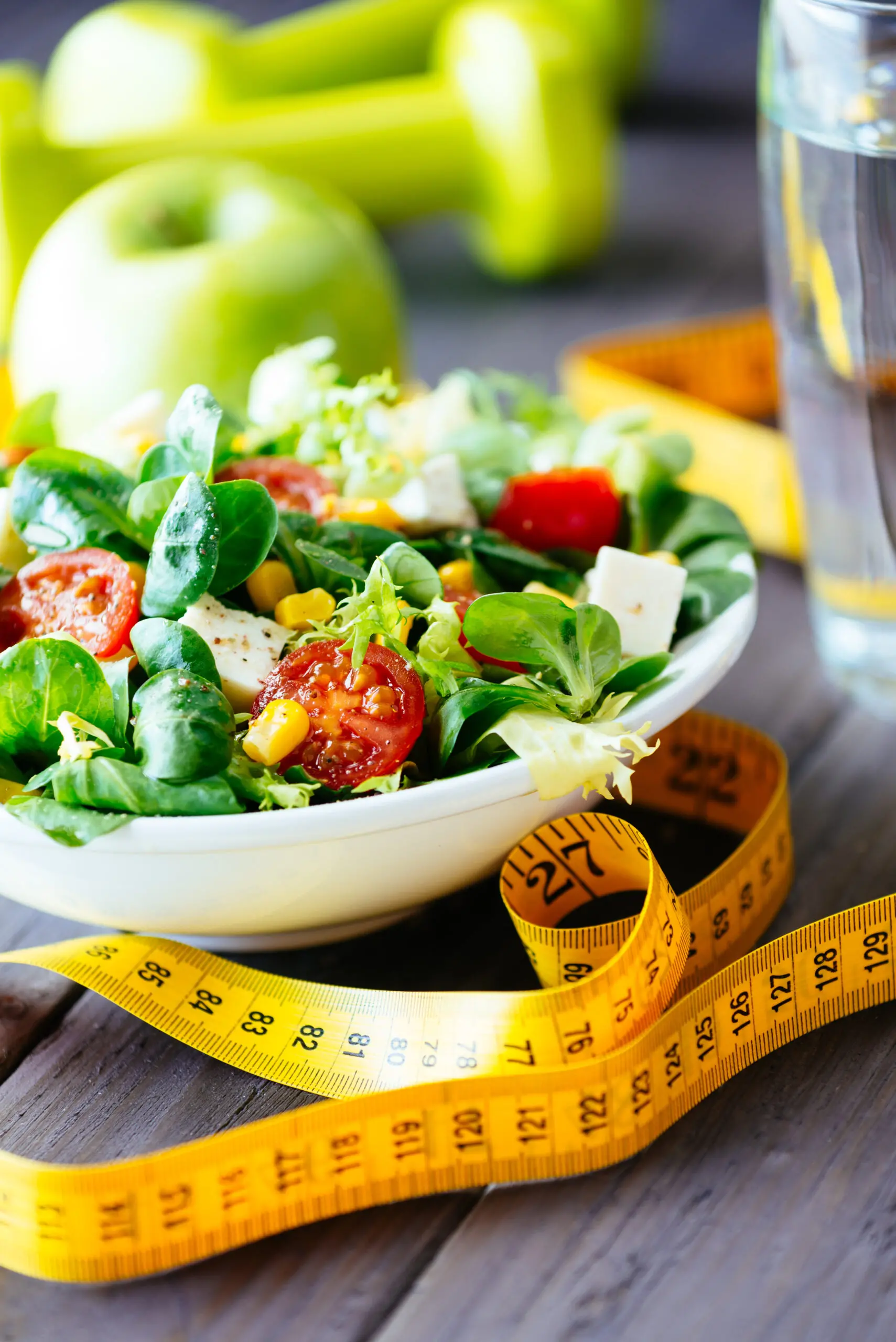 Bowl of fresh salad with cherry tomatoes, corn, and greens, a yellow measuring tape, green apples, dumbbells, and a glass of water on a wooden surface.