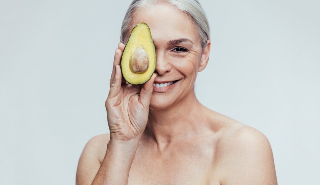 Smiling woman with light hair holding half an avocado over one eye against a plain light background