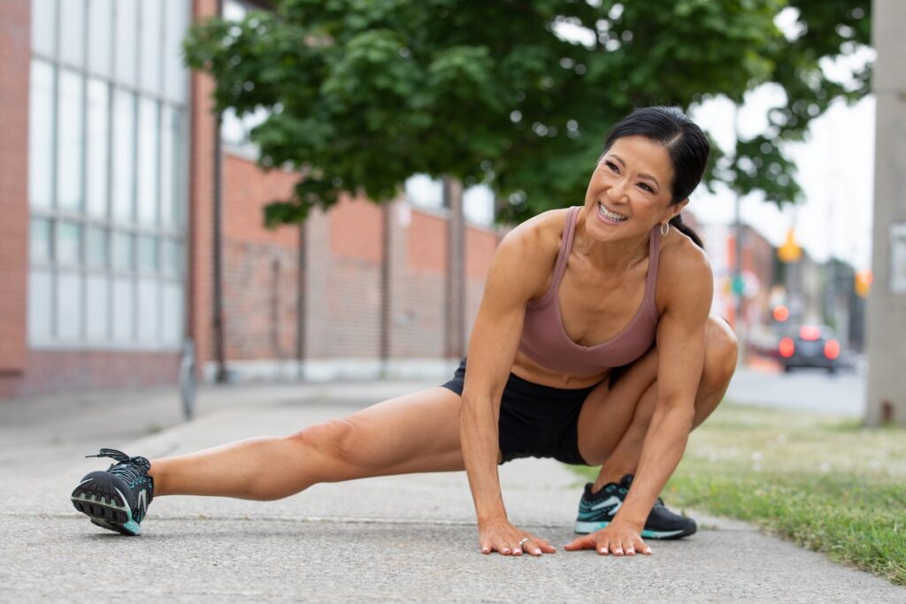 Woman in athletic wear stretching one leg out to the side on a sidewalk in an urban area, smiling and looking to the side.
