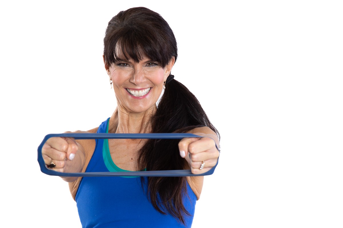 Smiling woman in blue tank top stretching a blue resistance band forward with both hands against a white background.
