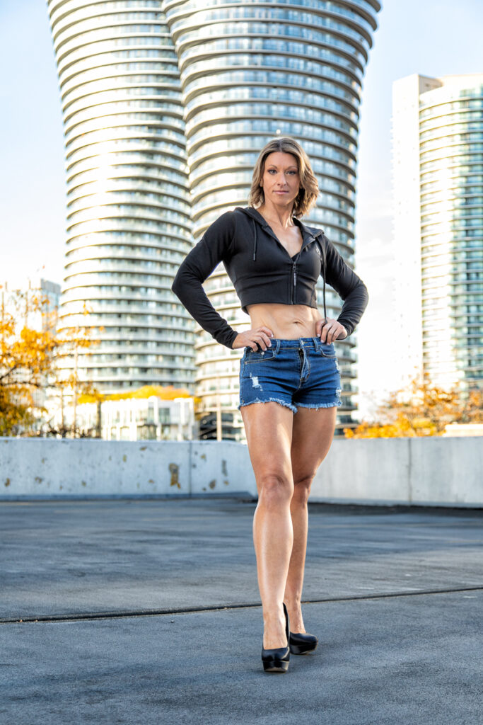 Woman in a cropped black jacket and jeans standing with her hands on her hips outside curved high-rise buildings.