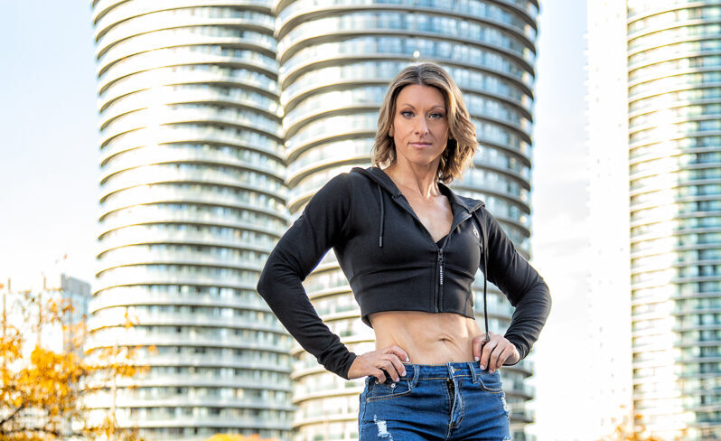 Woman in a cropped black jacket and jeans standing with her hands on her hips outside curved high-rise buildings.