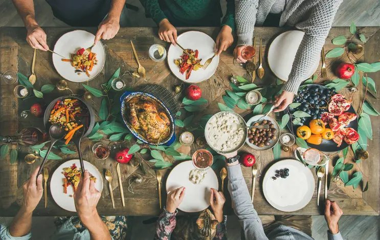 Overhead view of a wooden table set with plates of food, a roasted chicken, fruits, and people reaching for food with forks and spoons.
