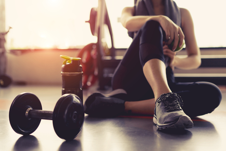 Person seated on a gym floor beside dumbbells in warm sunlight.