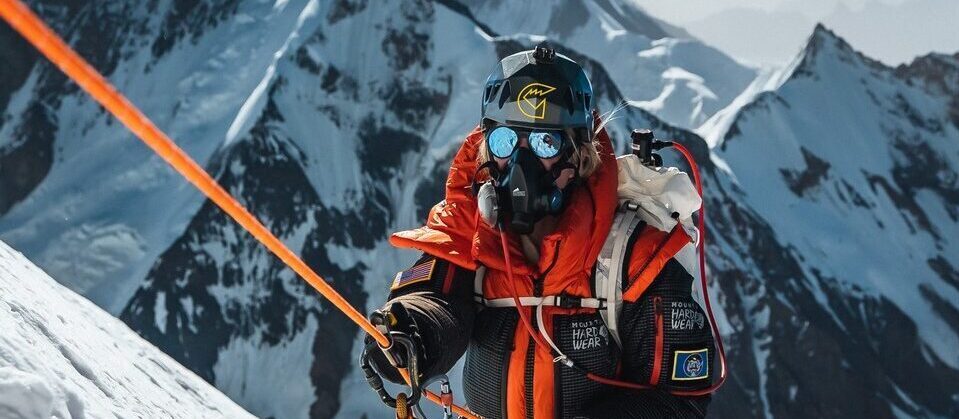 Mountaineer in an orange jacket and protective face gear on a snowy mountain beside a climbing rope.