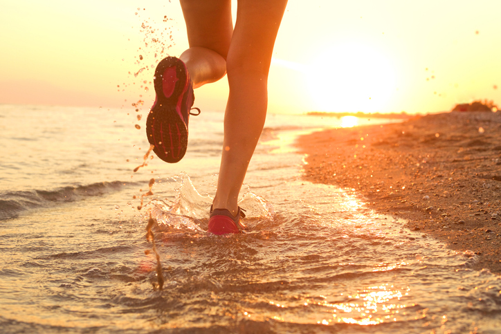 Close-up of a runner’s legs splashing through shallow water on a beach at sunset.