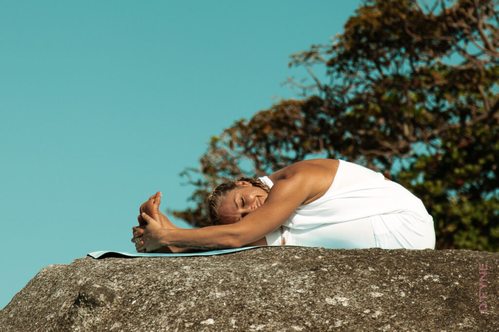 Woman folding forward over her extended legs while seated on a rock outdoors.