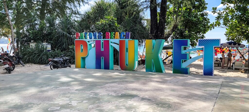 Colorful large letters spelling PHUKET with smaller letters above spelling PATONG BEACH, set on a beach with trees, motorcycles, and people under umbrellas in the background.