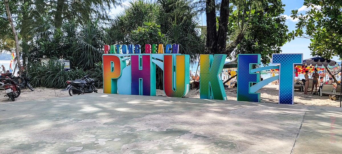 Colorful large letters spelling PHUKET with smaller letters above spelling PATONG BEACH, set on a beach with trees, motorcycles, and people under umbrellas in the background.