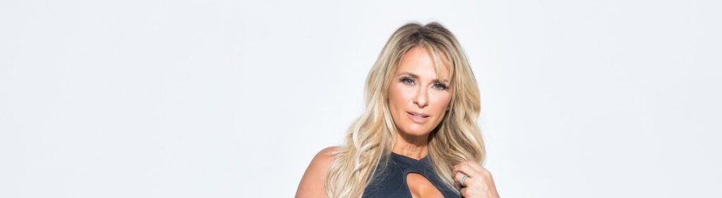 Blonde woman with long hair wearing a sleeveless black top, posing against a plain white background.