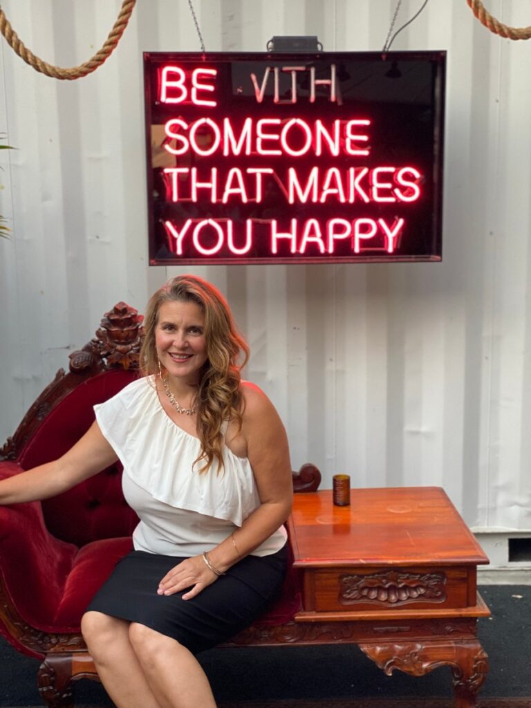 Woman in a white one-shoulder top and black skirt sitting on a red velvet vintage sofa next to a wooden table under a neon sign that reads 'BE WITH SOMEONE THAT MAKES YOU HAPPY'.