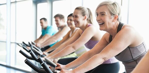 Five people smiling and exercising on stationary bikes in a bright gym.