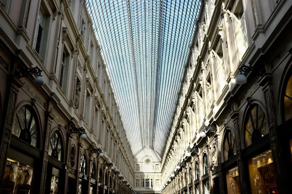 Interior view of a long, elegant shopping arcade with a high glass ceiling and ornate architectural details on both sides.