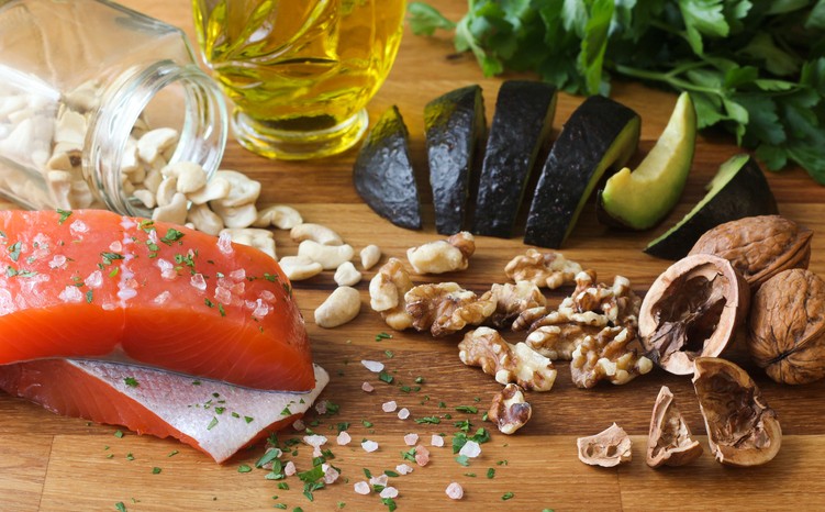 Two raw salmon fillets, sliced avocado, walnuts, cashews, a jar of cashews, a bottle of olive oil, and fresh herbs on a wooden surface.