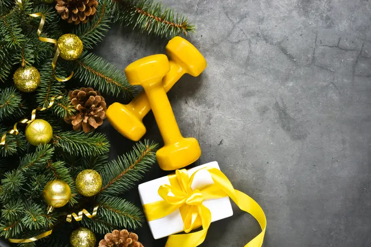 Yellow dumbbells, a white gift box with a yellow ribbon, pine cones, gold Christmas ornaments, and evergreen branches on a gray surface.