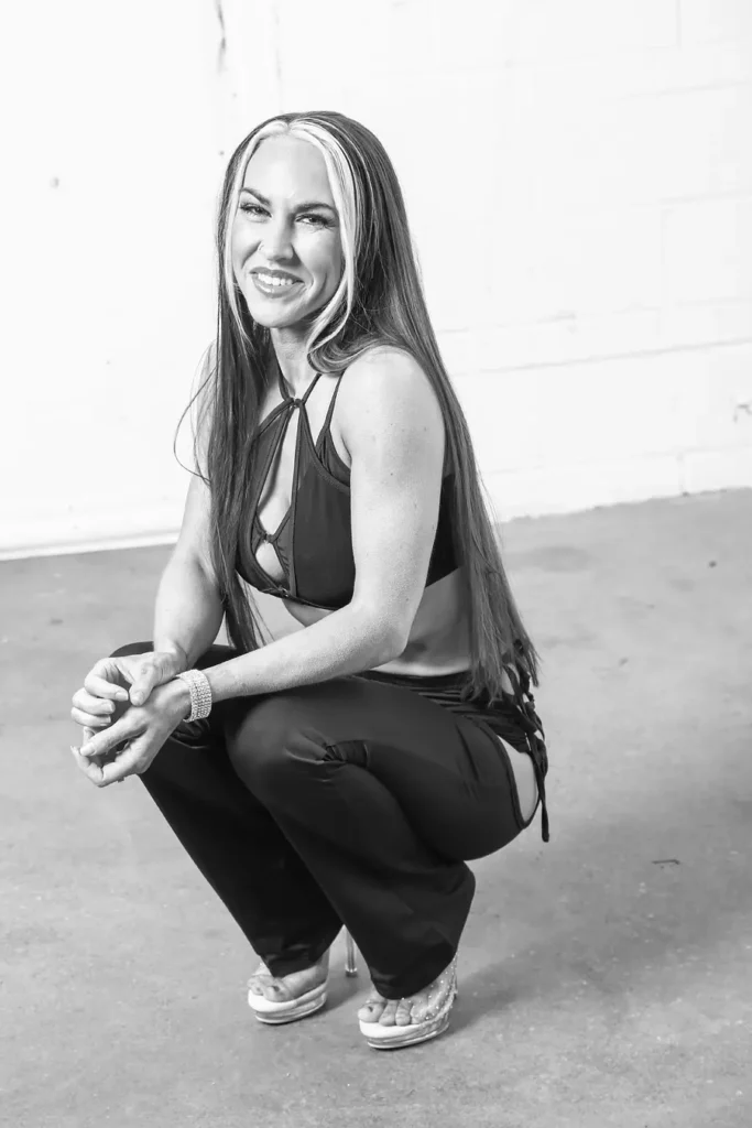 Woman with long hair featuring a light streak, wearing a sleeveless top and pants, squatting and smiling at the camera in a bright indoor space.