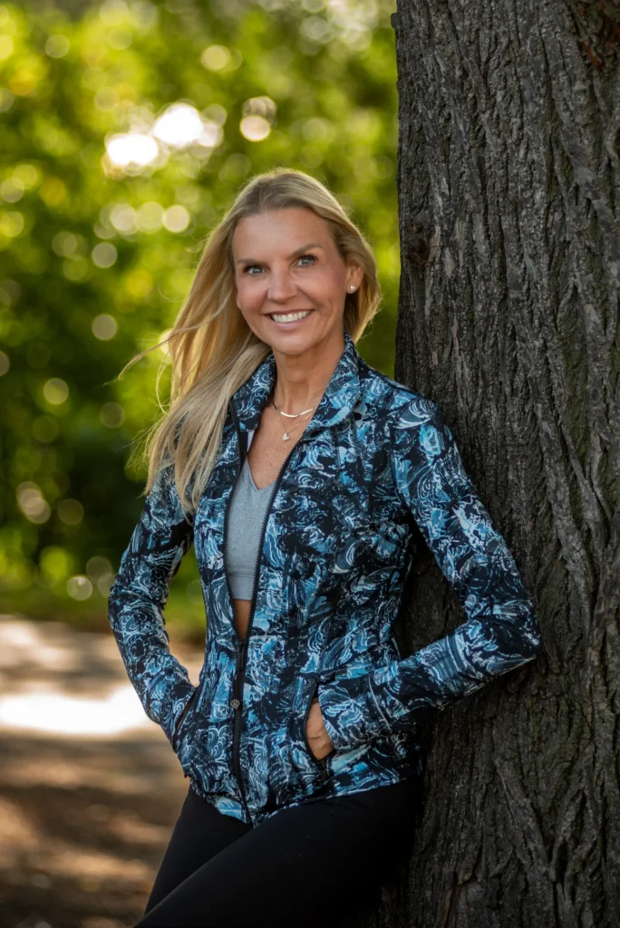 Smiling woman with long blonde hair wearing a blue patterned jacket and gray top, leaning against a tree with a blurred green background.