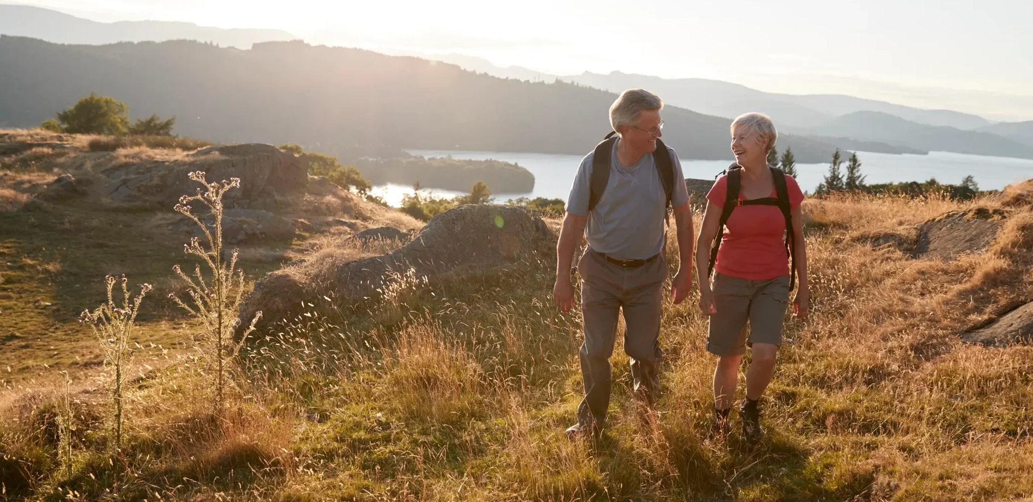 An older couple hiking on a grassy trail with backpacks, smiling at each other, with hills and a lake in the background at sunset.