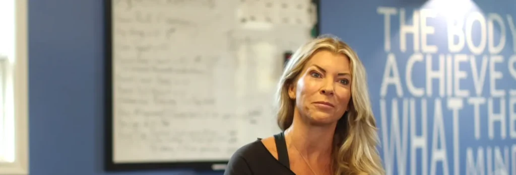 Blonde woman with long hair sitting indoors in front of a blue wall with white text and a blurred whiteboard.