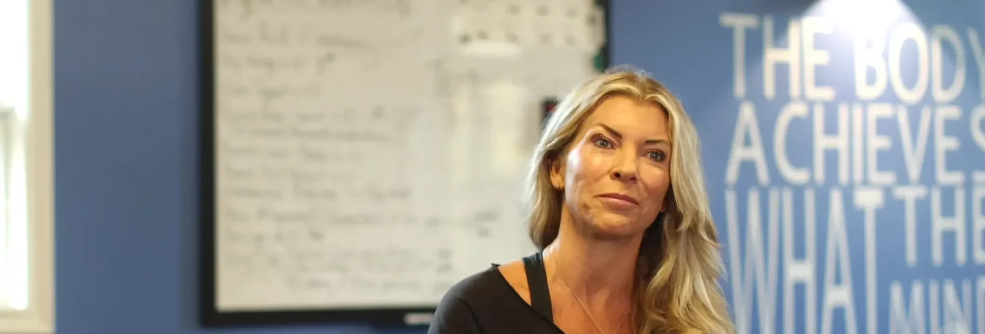 Blonde woman with long hair sitting indoors in front of a blue wall with white text and a blurred whiteboard.