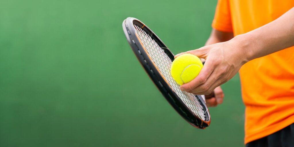 Person in orange shirt holding a tennis racket and a tennis ball against a green background.
