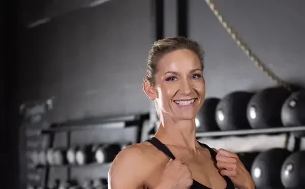 Smiling woman in workout attire standing in a gym with kettlebells and gym equipment in the background.