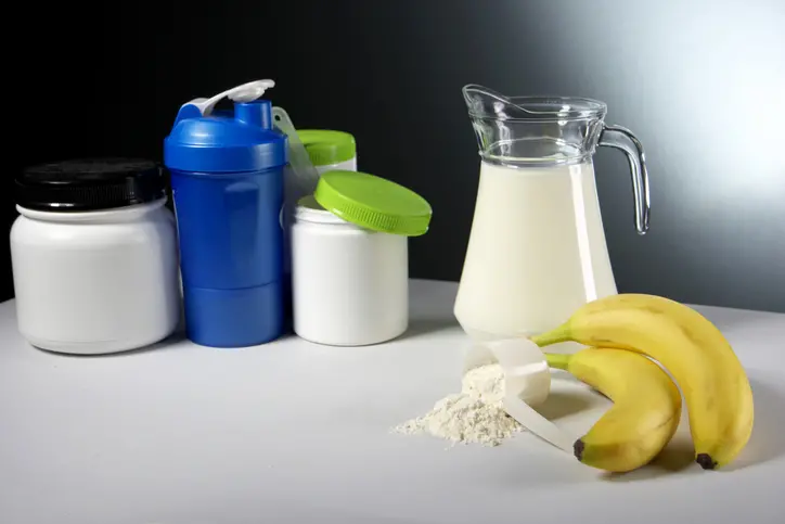 Various white supplement containers with green and black lids, a blue shaker bottle, a glass pitcher of milk, two bananas, and a scoop of white powder on a white surface.