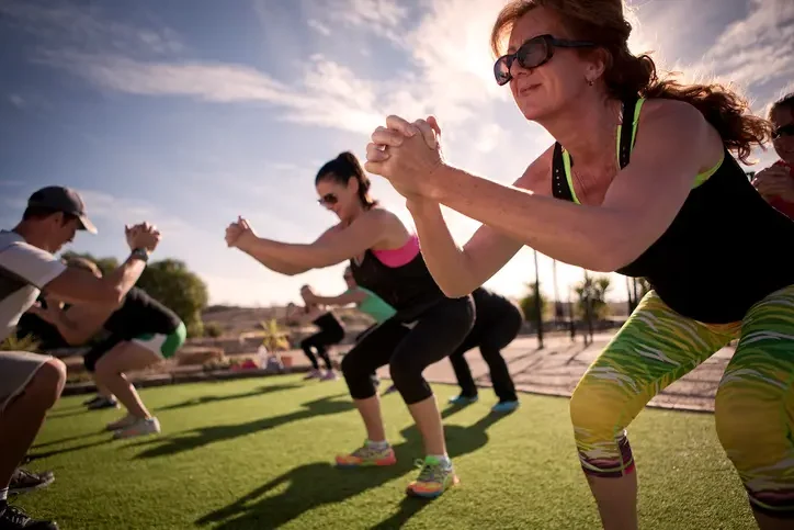 Group of adults exercising outdoors on grass, performing squats with hands clasped, wearing athletic clothing and sunglasses in sunny weather.