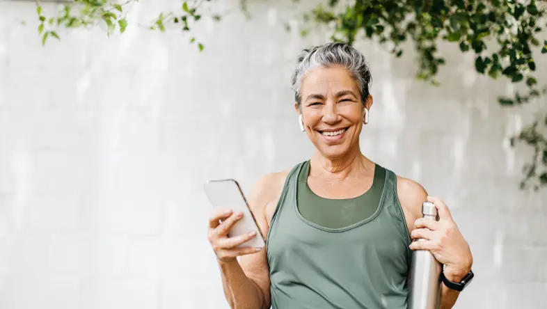 Smiling woman in workout clothes holding a smartphone and a water bottle, standing outdoors with greenery in the background.