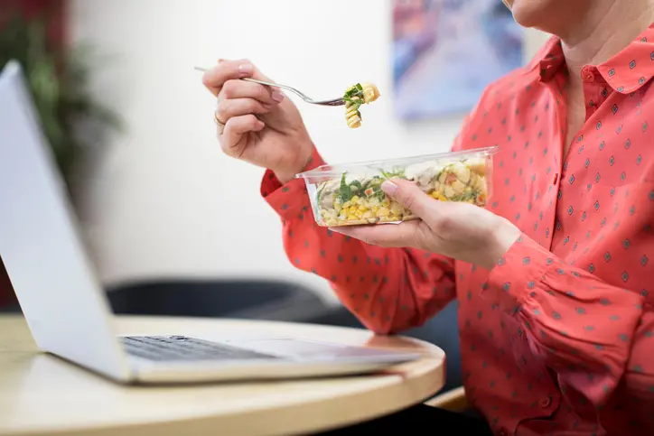 Person in a red patterned shirt eating a salad with pasta and vegetables from a clear container while sitting at a table with an open laptop.