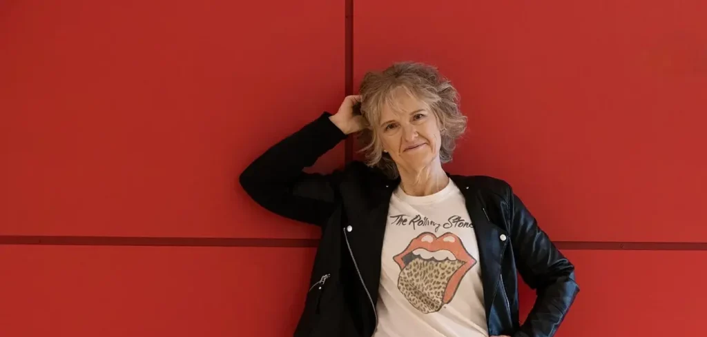 Person with short curly hair wearing a black jacket and a white Rolling Stones t-shirt, standing against a red wall with one hand on their head.