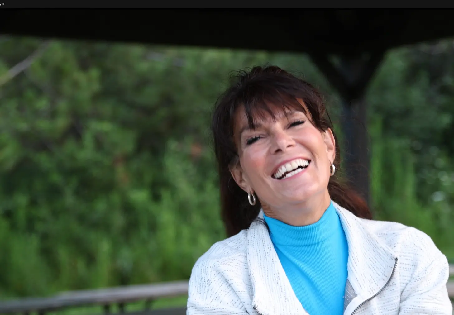 Smiling woman with dark hair wearing a blue turtleneck and light jacket outdoors with green foliage in the background.