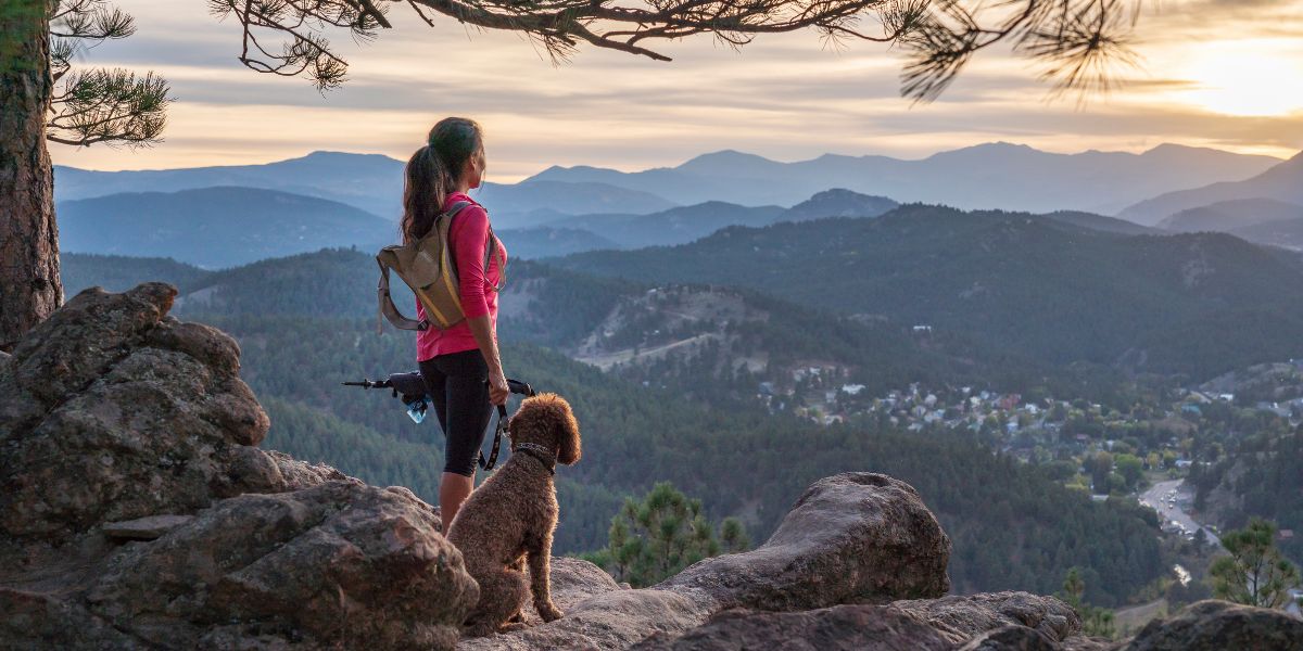 A woman with a backpack and her dog stand on a rocky overlook, gazing at a forested valley and mountain range at sunset.