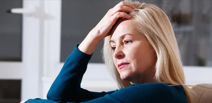 A middle-aged woman with blonde hair wearing a blue sweater sits with her hand on her forehead, looking thoughtful or stressed.