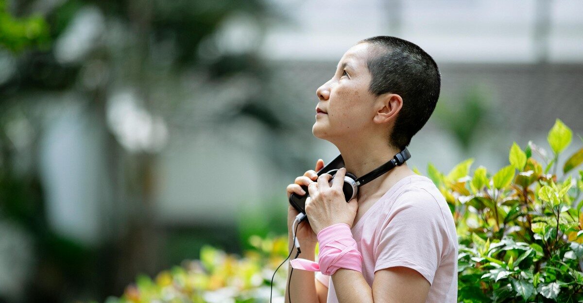 Person with short hair holding headphones around their neck, looking upward, standing outdoors near green plants.
